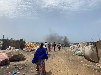 A group of people in a place with a lot of waste, smoke in the background. 